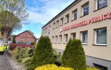 Dzieci w ż&oacute;łtych kamizelkach odblaskowych ze swoimi Paniami stoją przed biblioteką w Zblewie.