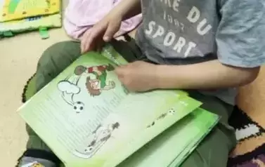 Chlopiec w ciemnych włosach i siwej koszulce siedzi na dywanie i przegląda książkę z bajkami.