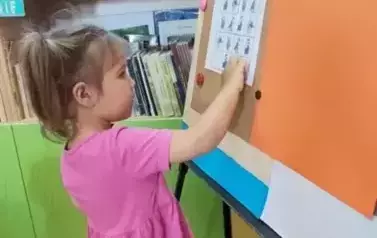Dziewczynka w r&oacute;żowej sukience przyczepia na metalową tablicę kolejną część układanki sudoku o świnkach.