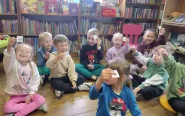 Dzieci siedzą na siedziskach i mają podniesione ręce w g&oacute;rze z karteczką ze znaczkiem &quot;dobrze&quot;