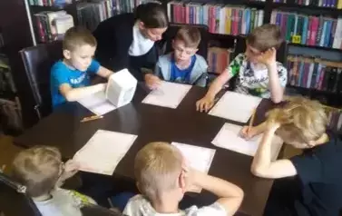 Chłopcy przy stole wypełniają karty pracy. Ich Pani pochyla się nad stołem.