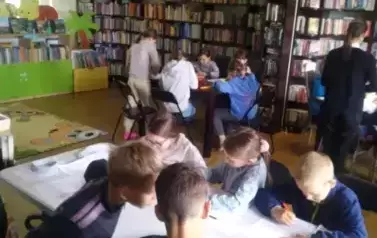 Dzieci siedzą przy stołach i wypełniają karty pracy. Przy jednym ze stolik&oacute;w stoi nauczycielka.