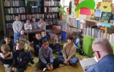 Dzieci siedzą przy stoliku i na dywanie i słuchają bajki, ktorą czyta im gościnnie kierowca przewoz&oacute;w szkolnych Pan Wojciech Formela