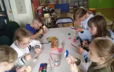 Dzieci przy stole malują farbami drewniane kwiatki na Dzień Matki.