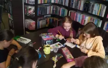 Dziewczynki przy stole malują drewniane kwiatki na Dzien Matki. Na stole są  pisaki, kredki i farby.