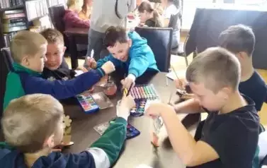 Dzieci siedzą przy stole malują farbami i pisakami drewniane kwiatki na Dzień Matki. Przy stole stoi Nikola Sturmowska.