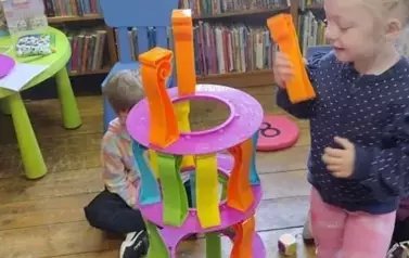 Dziewczynka w blond wlosach stoi przy wieży z klock&oacute;w trzymając w ręku klocek plastikowy.