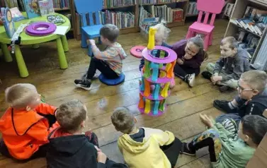 Dzieci siedzą w k&oacute;łeczku, a po środku jest ułożona wieża z plastikowych klock&oacute;w.