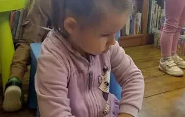 Dziewczynka w kitku siedzi na podłodze i bawi się pluszową książeczką. W tle widać inne dzieci.