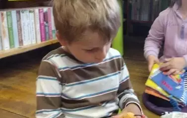 Chłopiec w blond włosach bawi się książeczką w kształcie ślimaka. Obok widać dziewczynkę, kt&oacute;ra też się bawi pluszową książeczką.