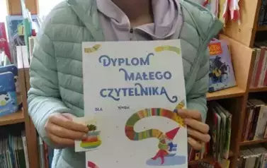 Dziewczynka w blond włosach i w seledynowej kurtce stoi w bibliotece w Borzechowie i pokazuje sw&oacute;j otrzymany dyplom.