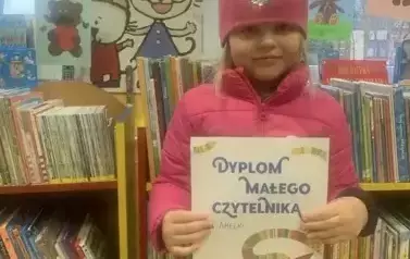 Dziewczynka w r&oacute;żowej kurtce i czapce stoi przy p&oacute;łkach z książkami w bibliotece w Zblewie i pokazuje sw&oacute;j dyplom.