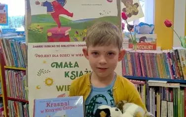 Chłopiec w blond włosach stoi przy tablicy z plakatem dotyczącym akcji Mała Książka Wielki Człowiek. Trzyma w ręku nową książkę i dwa pluszowe pieski.