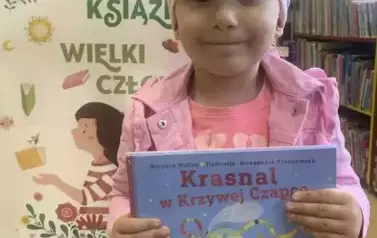 Dziewczynka ubrana na r&oacute;żowo stoi przy tablicy z plakatem dotyczącym akcji Mała Książka Wielki Człowiek. Pokazuje swoją nową książkę z wyprawki.