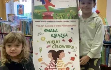 Przy tablicy z plakatami dotyczącymi akcji Mała Książka Wielki Człowiek stoi po prawej stronie chłopiec, a po lewej stronie dziewczynka trzymając nową książkę.