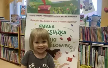Uśmiechnięta dziewczynka w blond włosach stoi przy tablicy z plakatami dotyczącymi akcji Mała Książka Wielki Człowiek, trzyma w rączkach nową książkę.