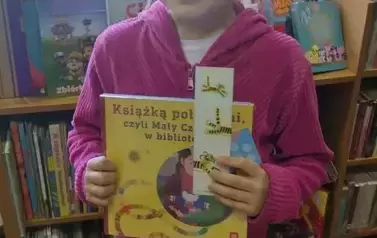 Dziewczynka w ciemnych wlosach i r&oacute;żowej bluzie stoi w bibliotece w Borzechowie pokazując swoją nową książkę i zakładkę do książki.