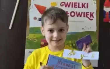 Chłopiec w blond włosach stoi przy drzwiach na kt&oacute;rych wisi plakat Mała Książka Wielki Człowiek. Trzyma w ręku nową książkę, zakładkę i kartę Małego Czytelnika.