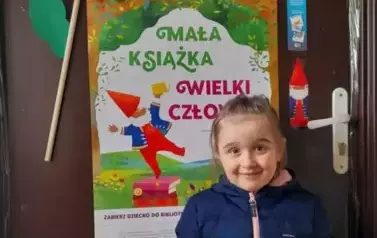 Dziewczynka w jasnych włosach i w granatowej kurtce stoi przy brązowych drzwaich na kt&oacute;rych wisi plakat Mała Książka Wielki Człowiek i pokazuje swoją nową książkę.