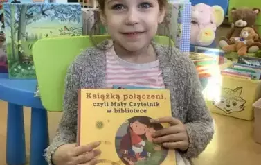 Dziewczynka w blond włosach i w siwym sweterku siedzi na zielonym krzesełku i pokazuje swoją nową książkę.