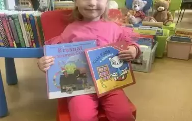 Dziewczynka w blond włosach i r&oacute;żowej bluzce siedzi na czerwonym krzesełku i pokazuje nową książkę i książkę, kt&oacute;rą wypożyczyła. Uśmiecha się.