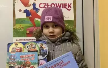 Dziewczynka w siwej kurtce i fioletowej czapce stoi przy drzwiach na kt&oacute;rych wisi plakat akcji Mała Książka Wielki Człowiek. Trzyma nową książkę i książkę wypożyczoną.