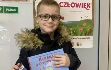 Chłopiec w blond włosach w okularach i ciemnej kurtce stoi przy drzwiach na kt&oacute;rych jest plakat Mała Książka Wielki Człowiek. Trzyma nową książkę i kartę Małego Czytelnika.