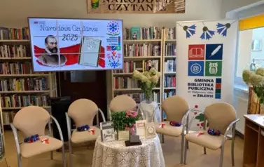 Przygotowane miejsce na Narodowe Czytanie. Pięć krzeseł na kt&oacute;rych są wianki i kotyliony. Po środku stoi stolik okrągły z książkami Jana Kochanowsiego. Wyżej jest duży ekran z wyświetlanym plakatem z tego wydarzenia, po prawej stoi rolap z logiem bibliote