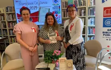 Przy okrągłym stoliku stoi szefowa biblioteki z dwiema Paniami: z Panią Radną Gminy i z Panią Sekretarz Gminy. Mają przyczepione kotyliony i wianki na głowach. Kierowniczka stojąca w środku trzyma książkę z poezją Jana Kochanowskiego.