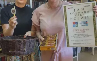 Bibliotekarka stoi z Panią Radną Gminy, kt&oacute;ra trzyma plakat z wydarzenia i karton z wafelkami, bibliotekarka trzyma koszyk z zapinkami i długopisami, jeden długopis z zapinką trzyma  w ręce pokazując go. 