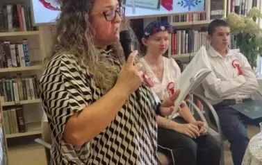 Kierownik Biblioteki czyta tren Jana Kochanowskiego, czytająca młodzież siedzi przy stoliku, nad głowami wisi duży ekran z wyświetlanym plakatem tego wydarzenia.
