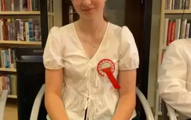 Na krześle siedzi dziewczynka z wiankiem na głowie i przyczepionym kotylionem na piersi. Trzyma zieloną teczkę z tekstem do czytania .