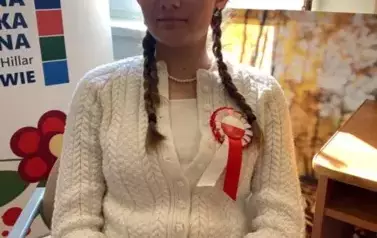 Na krześle siedzi dziewczyna w blond warkoczach i z wiankiem na głowie, na lewej piersi ma przyczepiony kotylion. Na nogach położona jest teczka zielona z tekstem do czytania.