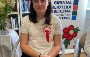 Na krześle siedzi dziewczyna w dłuższych ciemnych włosach w okularach i z wiankiem na głowie, na piersi ma przyczepione kotylion. Trzyma teczkę zieloną z tekstem do czytania .