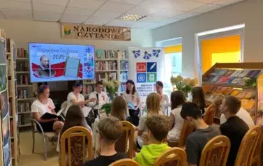 Młodzież siedzi na krzesłach i słucha jak ich kolega zaczyna czytać treny. Nad głowami jest duży ekran z wyświetlanym plakatem dot. wydarzenia,a po prawej stronie stoi rolap z logiem biblioteki