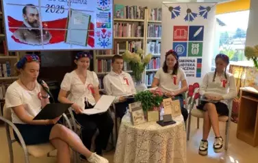 Przy okrągłym stoliku siedzą 4 dziewczyny i jeden chłopak, jedna z nich tzyma mikrofon i czyta na głos treny. Nad nimi jest duży ekran z wyświetlanym plakatem z wydarzenia.