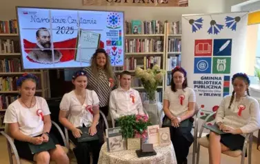 Przy okrągłym stoliku siedzą cztery dziewczyny i jeden chłopak, kt&oacute;rzy mają czytać poezje Jana Kochanowskiego. Za nimi stoi szefowa biblioteki, nad głowami wisi duży ekran z wyświetlanym plakatem z Narodowego Czytania.