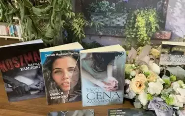 Nowe książki leżą na stole z widocznymi tytułami, po środku leży wianek z kwiat&oacute;w z książką w środku. W tle jest dom obrośnięty fioletowym bzem.