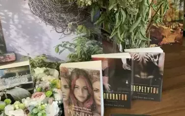 Nowe książki leżą na stole z widocznymi tytułami, po środku leży wianek z kwiat&oacute;w z książką w środku. W tle jest dom obrośnięty fioletowym bzem.
