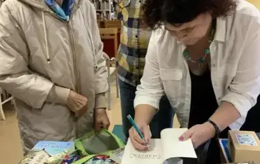 Przy wyłożonych książkach na biurku, pisarka podpisuje czytelniczce zakupioną książkę. Czytelniczka się bardzo uśmiecha, obok stoi mężczyzna z brodą, kt&oacute;ry jest mężem pisarki.