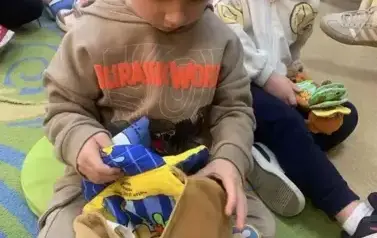 Chłopiec w blond włosach i beżowej bluzie siedzi na dywanie i bawi się pluszową misiową książeczką . Za nim widać inne dzieci bawiące się książeczkami.