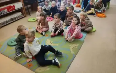 Dzieci siedzą na dywanie i siedziskach słuchając bajki, kt&oacute;rą czyta im bibliotekarka.