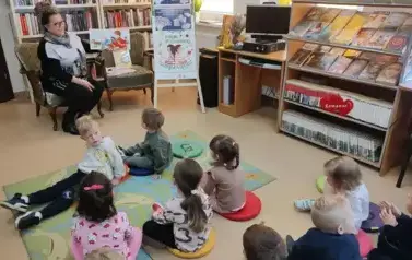 Dzieci siedzą na dywanie i siedziskach patrząc na bibliotekarkę, kt&oacute;ra ma obok na fotelu przygotowane bajeczki do czytania.
