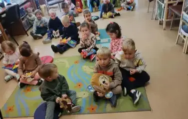 Dzieci siedzą na dywanie i siedziskach bawiąc się pluszowymi książeczkami.