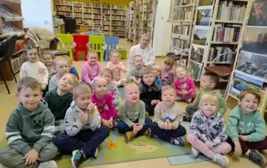 Dzieci siedzą na dywanie, za nimi siedzi ich Pani.