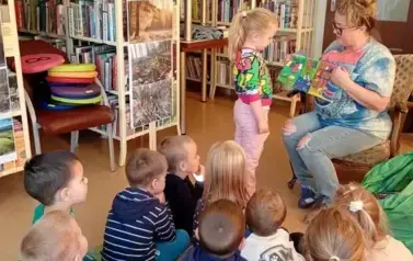 Dzieci siedzą na dywanie i patrzą na swoją koleżankę, kt&oacute;ra stoi przy bibliotekarce, kt&oacute;ra trzyma pluszową książeczkę przedstawiającą sklep, a dziewczynka ma robić z niego zakupy.
