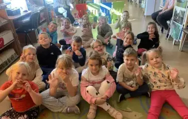 Dzieci siedzą na dywanie, niekt&oacute;re z nich mają podniesioną rękę w celu zabrania głosu.