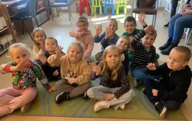 Dzieci siedzą na dywanie, niekt&oacute;re z nich mają podniesioną rękę w celu zabrania głosu.