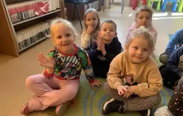 Dzieci siedzą na dywanie, jedna dziewczynka ma podniesioną lekko rączkę jakby chciała nią pomachać.