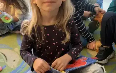 Dziewczynka w blond włosach trzyma pluszową książeczkę,  w tle widać inne dzieci.